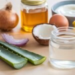 A photo of a table with various ingredients. There's a jar of aloe vera gel, a jar of coconut oil, a jar of honey, a sliced onion, a whole egg, a bowl of yogurt, and a lemon. The aloe vera gel, coconut oil, honey, and lemon are placed in a row. The sliced onion, whole egg, and bowl of yogurt are placed in another row. The aloe vera gel has a small amount of liquid inside. The background is light.