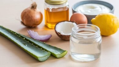 A photo of a table with various ingredients. There's a jar of aloe vera gel, a jar of coconut oil, a jar of honey, a sliced onion, a whole egg, a bowl of yogurt, and a lemon. The aloe vera gel, coconut oil, honey, and lemon are placed in a row. The sliced onion, whole egg, and bowl of yogurt are placed in another row. The aloe vera gel has a small amount of liquid inside. The background is light.